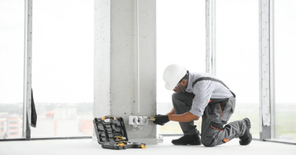 Een elektricien met witte helm, wit-zwart geruit hemd en grijze werkbroek met bretellen installeert een stopcontact in een nieuwbouwappartement. Daarvoor heeft de elektricien zijn kist met gereedschap nodig.